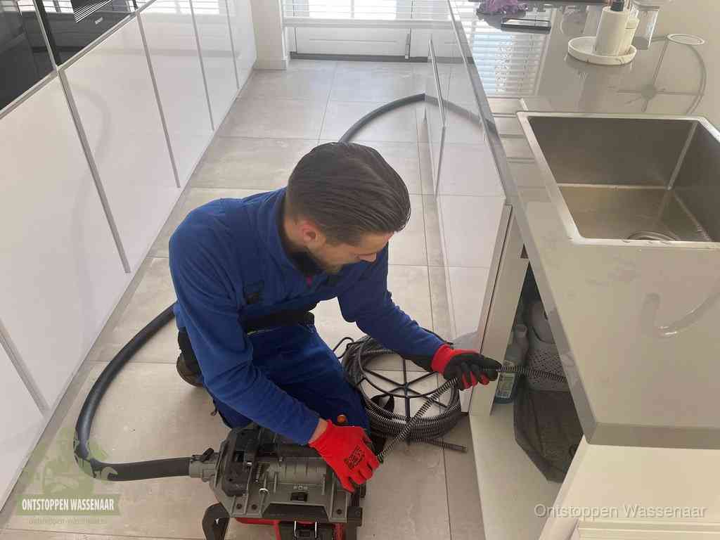 Een monteur ontstopt de keukenafvoer onder de spoelbak met een elektrische veermachine in Wassenaar