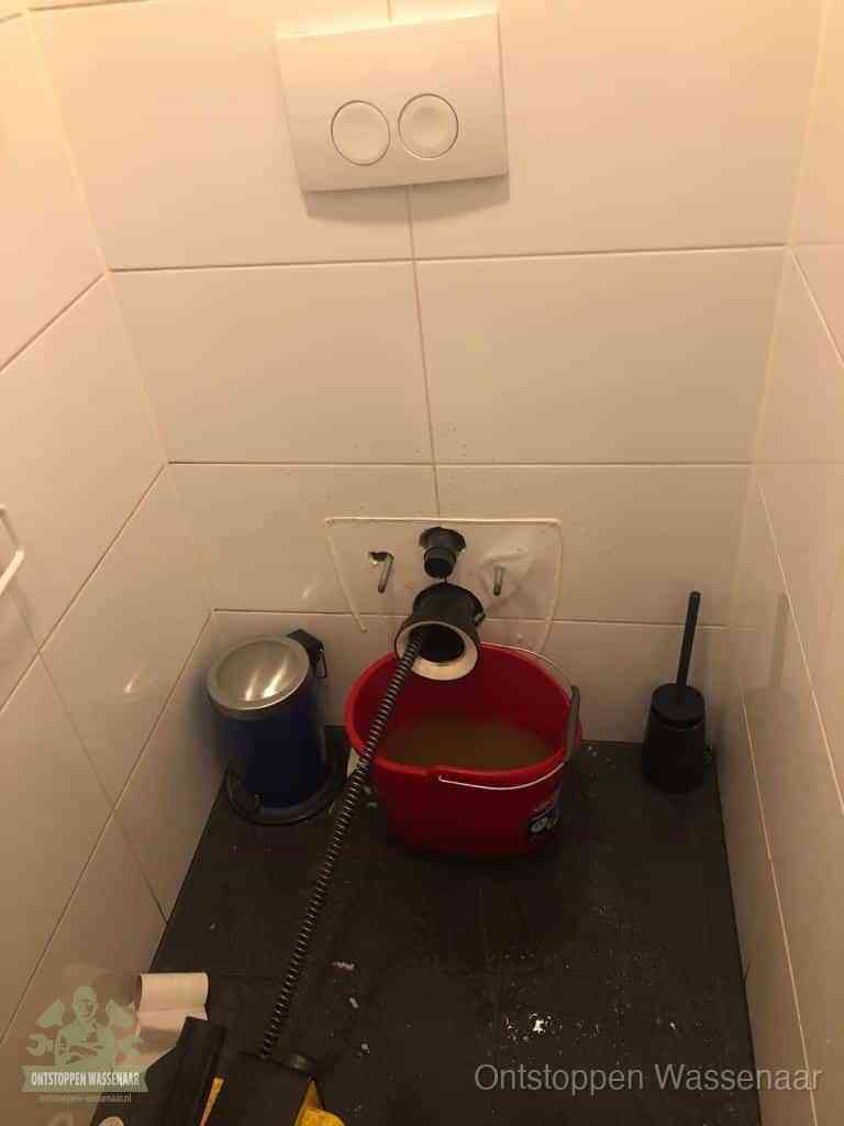 Een ontstopte wand-wc met verwijderde toiletpot waar een ontstoppingsveer in de wanduitgang verdwijnt, het achterhalen waar de verstopping zit in Wassenaar