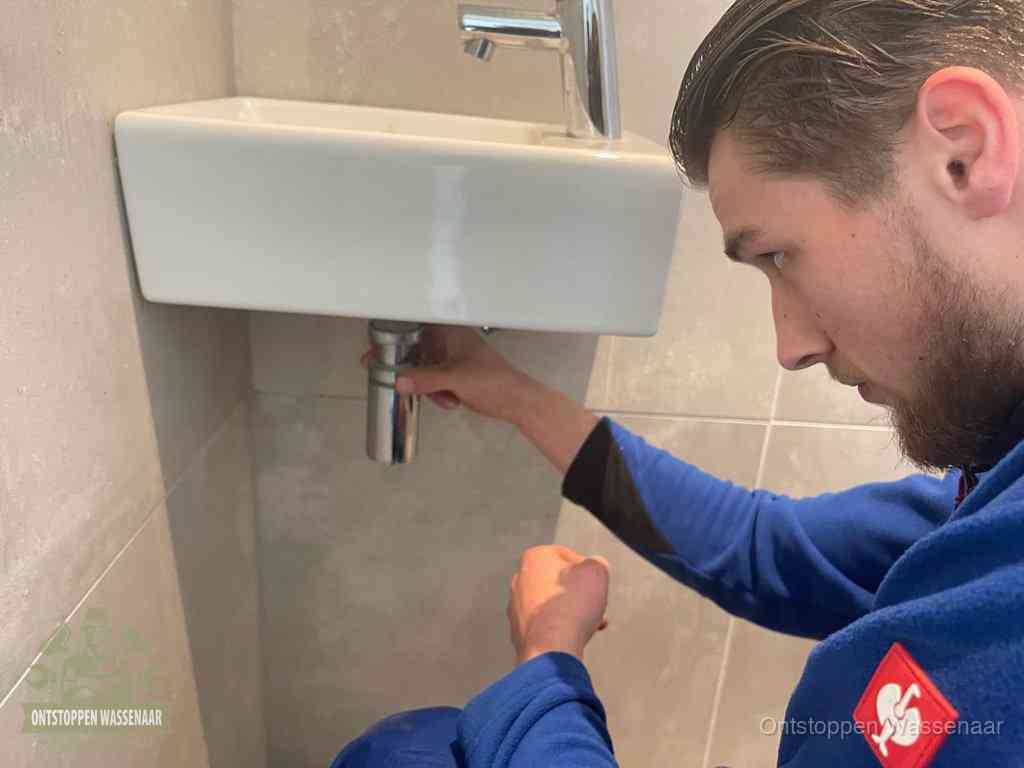 Een monteur draait met de hand de zwanenhalssifon onder een fonteinwasbak in de toiletruimte los om de bron van rioollucht in Wassenaar te vinden