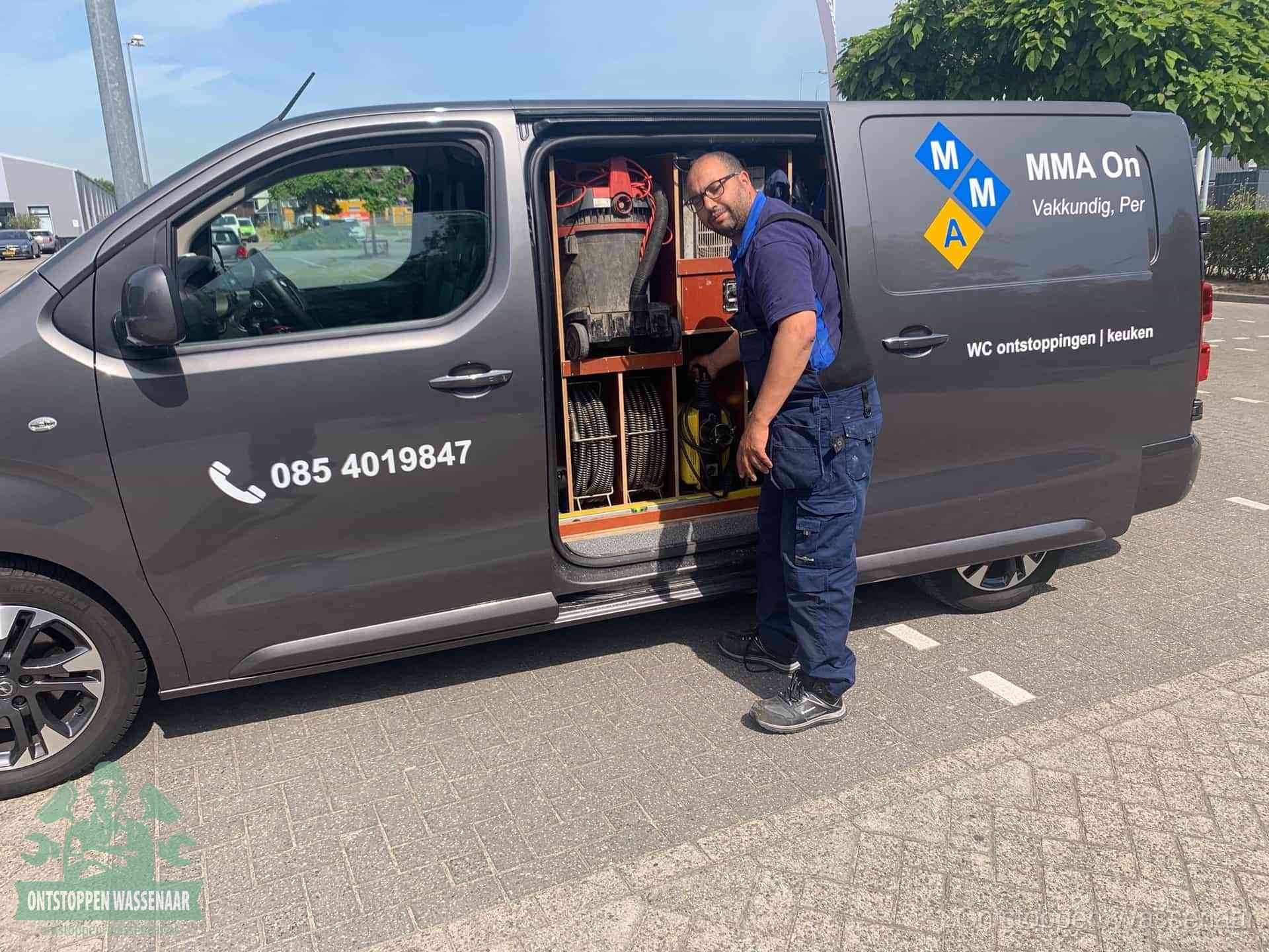 Een monteur bij de open schuifdeur van zijn bedrijfsbus op een parkeerplaats, klaar om de oorzaak van terugkerende afvoerverstoppingen aan te pakken in Wassenaar