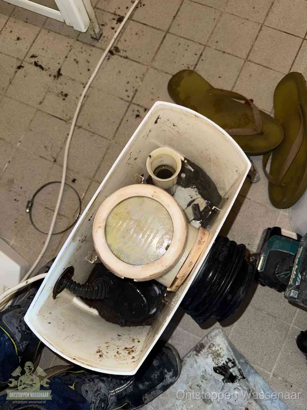 Een gedemonteerde toiletpot op de vloer met ontstoppingsveer en gereedschap ernaast, het verwijderen van een vastzittend voorwerp uit de wc in Wassenaar