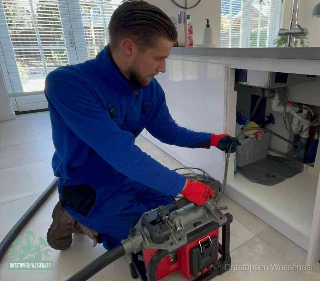 Een monteur met rode handschoenen knielt in een keuken en voert een elektrische ontstoppingsveer in tijdens het professioneel afvoer ontstoppen in Wassenaar