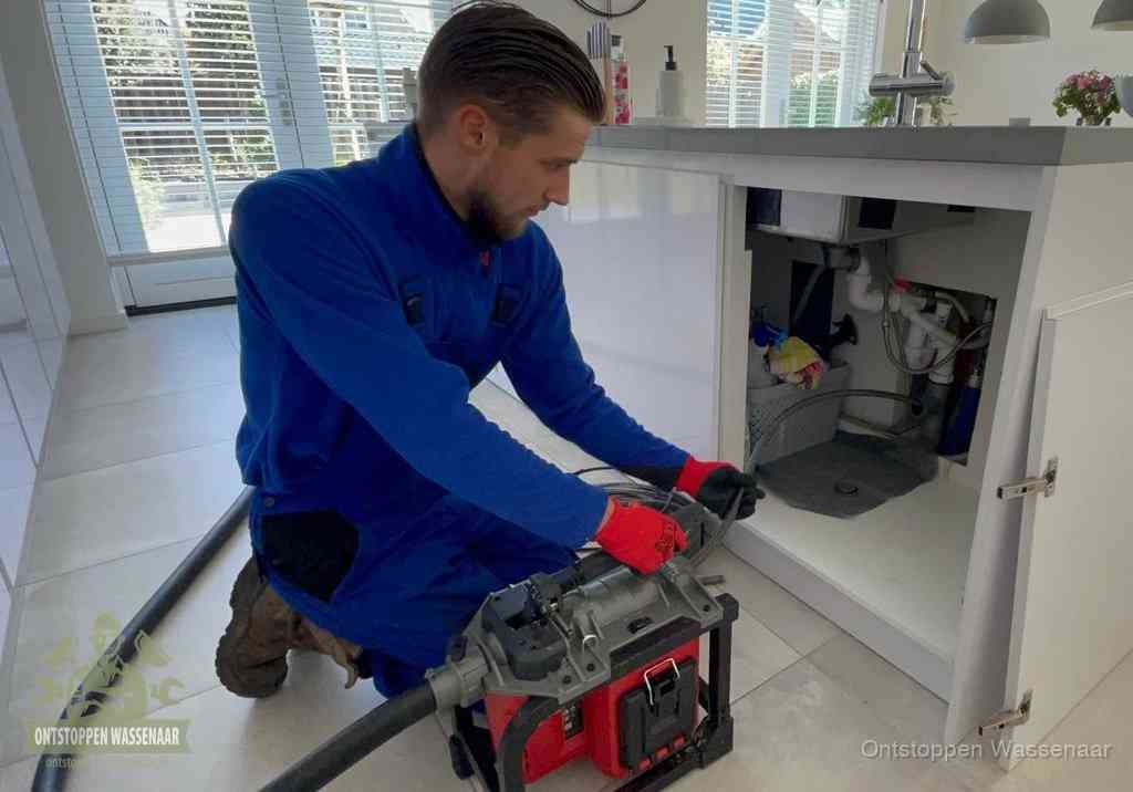 Een monteur voert een professionele elektrische ontstoppingsmachine in de afvoer onder de spoelbak tijdens het gootsteen ontstoppen in Wassenaar